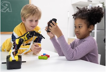 A young white boy and black girl sit at a classroom table looking at a small yellow and black table-top robot that the girl is controlling with a remote.
